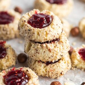 A stack of raspberry hazelnut thumbprint cookies filled with red jam, surrounded by more cookies on parchment paper.