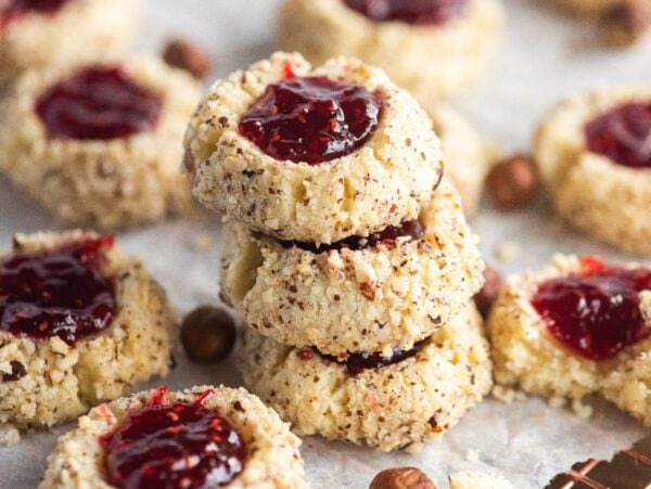 A stack of raspberry hazelnut thumbprint cookies filled with red jam, surrounded by more cookies on parchment paper.