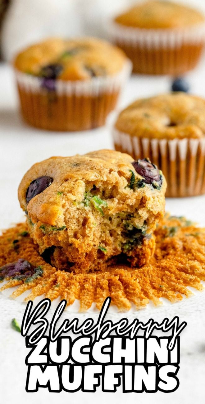 A blueberry zucchini muffin with a bite taken out, two whole muffins in the background. Text reads "Blueberry Zucchini Muffins"—a healthy blueberry muffin perfect for any zucchini muffin recipe.