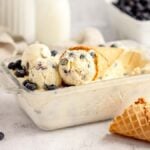 Glass dish of homemade cheesecake ice cream with a waffle cone, fresh blueberries, and a milk bottle in the background—made with fresh blueberries and cream cheese for a rich, tangy twist.