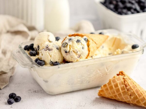 Glass dish of homemade cheesecake ice cream with a waffle cone, fresh blueberries, and a milk bottle in the background—made with fresh blueberries and cream cheese for a rich, tangy twist.