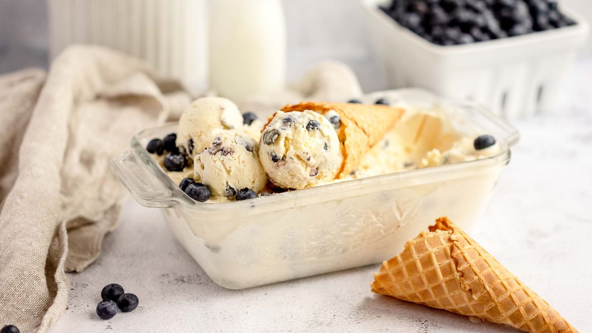 Glass dish of homemade cheesecake ice cream with a waffle cone, fresh blueberries, and a milk bottle in the background—made with fresh blueberries and cream cheese for a rich, tangy twist.