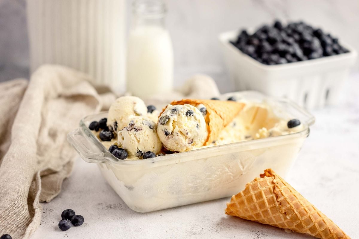 Glass dish with Blueberry Cheesecake Ice Cream, a waffle cone, fresh blueberries, and a bottle of milk in the background—made with fresh blueberries and cream cheese for a luscious treat.