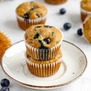 Two healthy blueberry muffins stacked on a plate, with more muffins and scattered blueberries in the background—perfect for anyone craving a delicious zucchini muffin recipe.