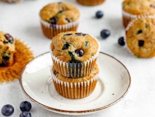 Two healthy blueberry muffins stacked on a plate, with more muffins and scattered blueberries in the background—perfect for anyone craving a delicious zucchini muffin recipe.