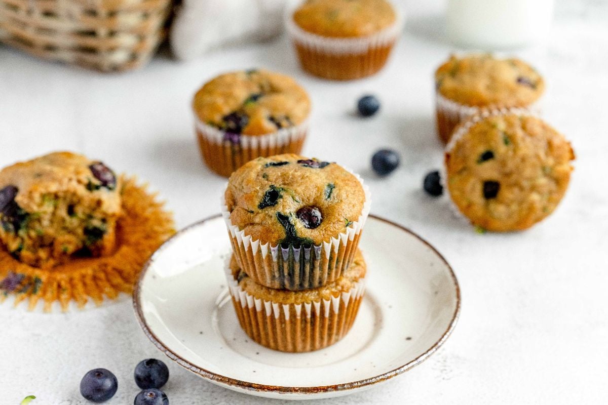 Two blueberry muffins stacked on a plate, with more muffins and scattered blueberries in the background—an inviting display perfect for anyone craving a healthy blueberry muffin.