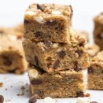 Stacked blonde brownies with chocolate chips and nuts rest on a white surface, with more blondie bars in the background.