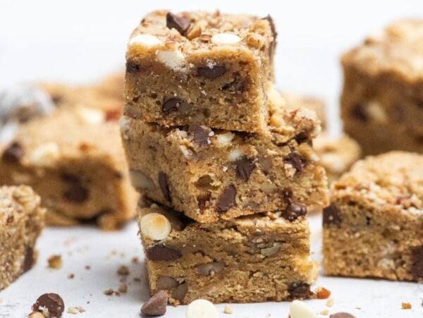 Stacked blonde brownies with chocolate chips and nuts rest on a white surface, with more blondie bars in the background.