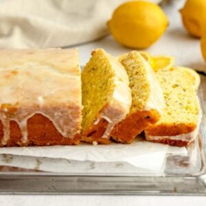 Iced lemon zucchini bread with several slices cut, lemons and lemon wedges in the background on a light surface.