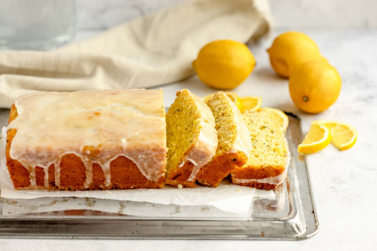 A glazed lemon zucchini bread loaf, sliced, with whole and sliced lemons in the background on a metal tray.
