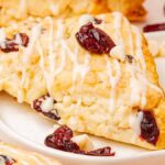 A delightful white chocolate cranberry scone is topped with white icing and dried cranberries on a plate, surrounded by creamy white chocolate chips.
