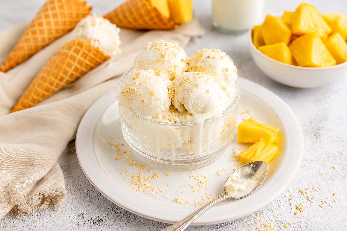 A glass bowl of frozen yogurt ice cream with coconut flakes, surrounded by pineapple chunks and ice cream cones—a Piña Colada frozen treat.