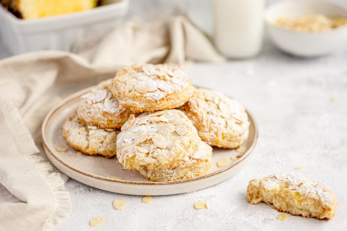 A plate of almond cookies dusted with powdered sugar, with a bitten cookie and milk in the background—perfect alongside pineapple scones made from the best fruit scone recipe.