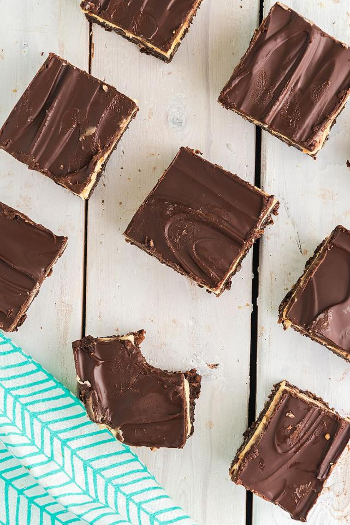 Chocolate-topped Nanaimo bars—an irresistible no bake dessert—sit on a white wooden surface near a teal napkin, one with a bite taken.