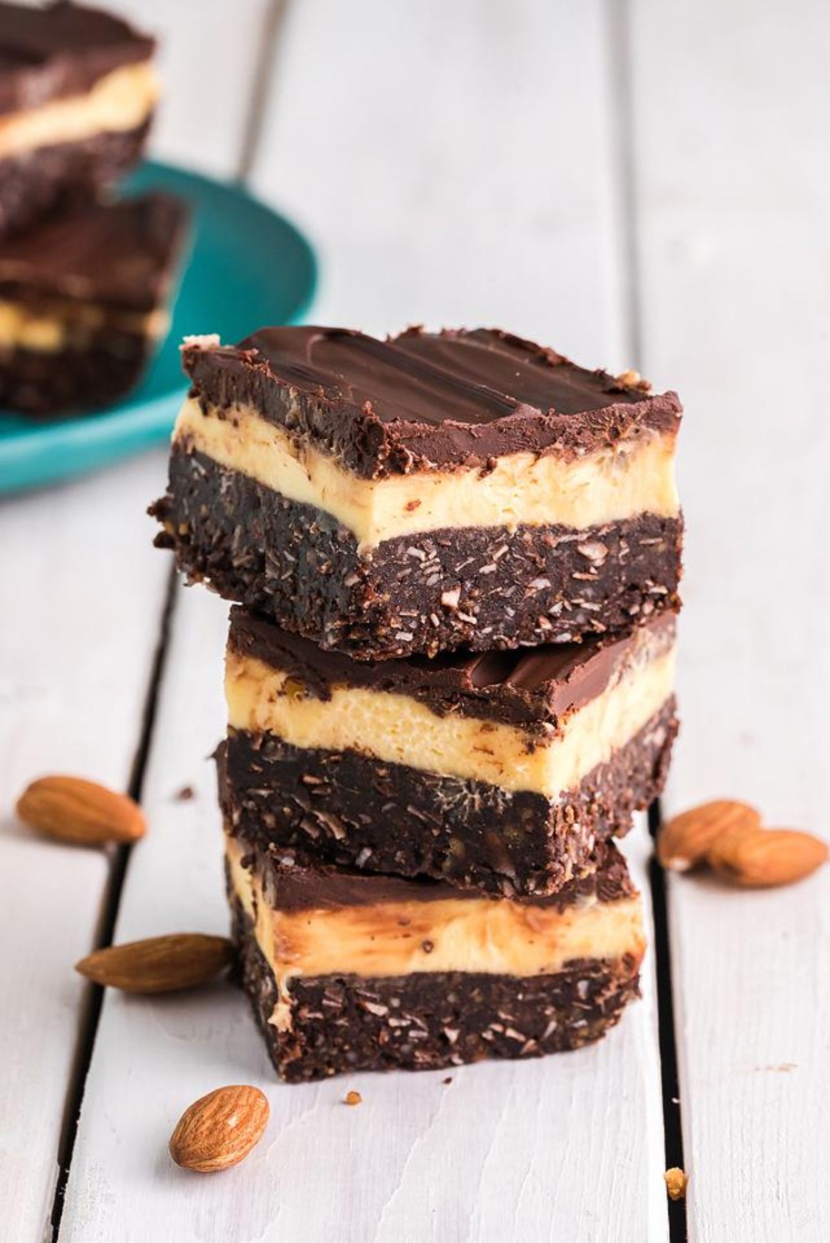 Three layered chocolate and cream bars, a classic Canadian dessert, stacked on a white wooden surface with almonds scattered around.