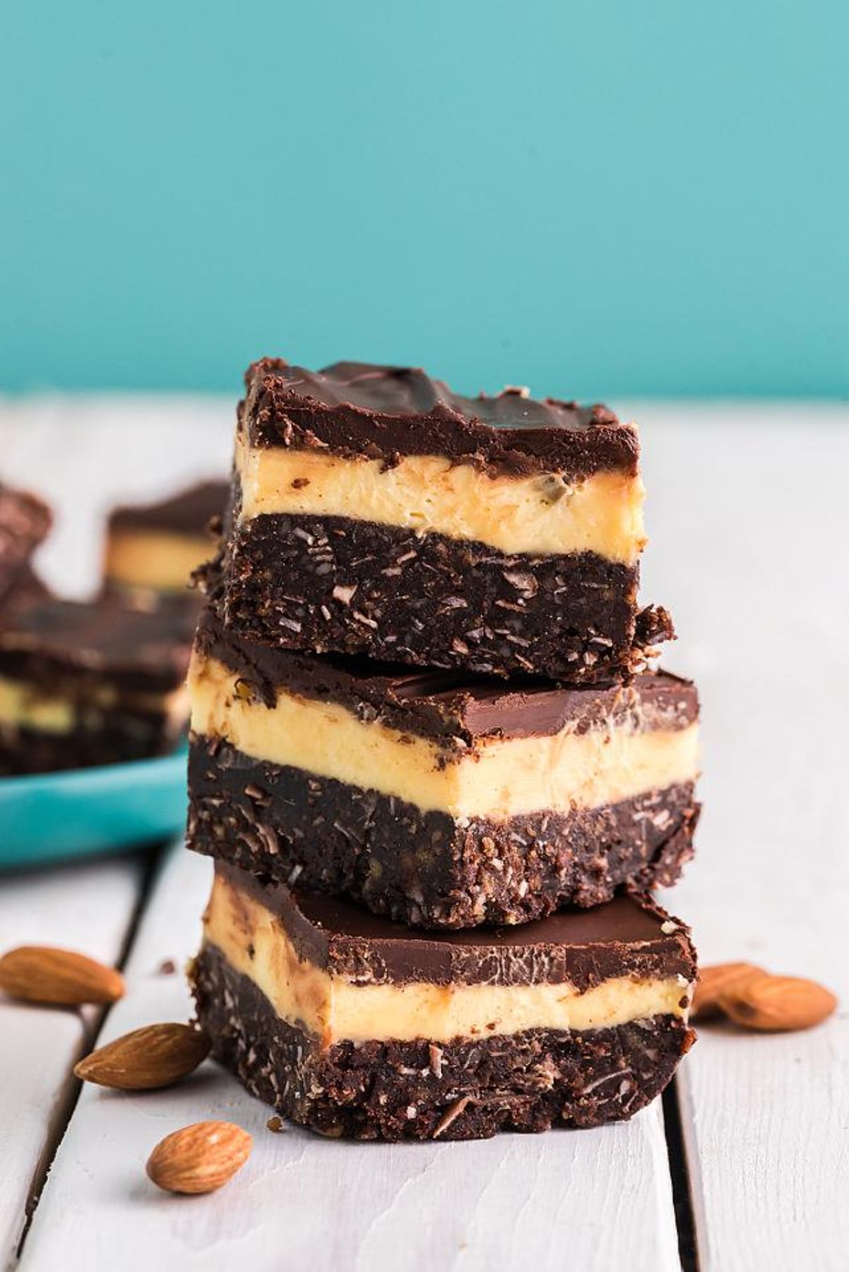 Three stacked Nanaimo bars with chocolate, custard, and coconut layers sit on a white surface with almonds nearby—an irresistible no bake dessert that’s perfect for any layered bar recipe collection.