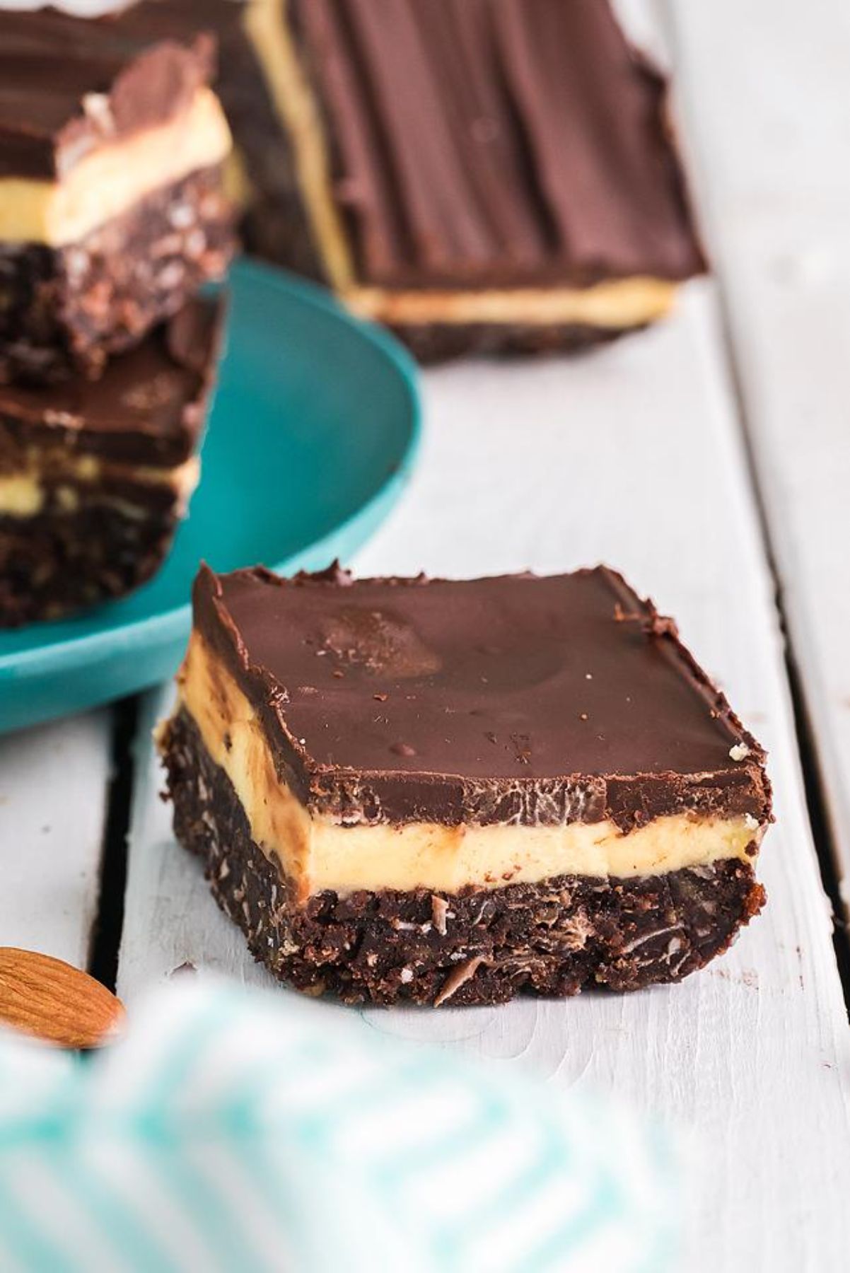 A square Nanaimo bar with chocolate, custard, and coconut layers—a classic Canadian dessert—sits on a white wooden surface. These indulgent bars freeze well for make-ahead treats.