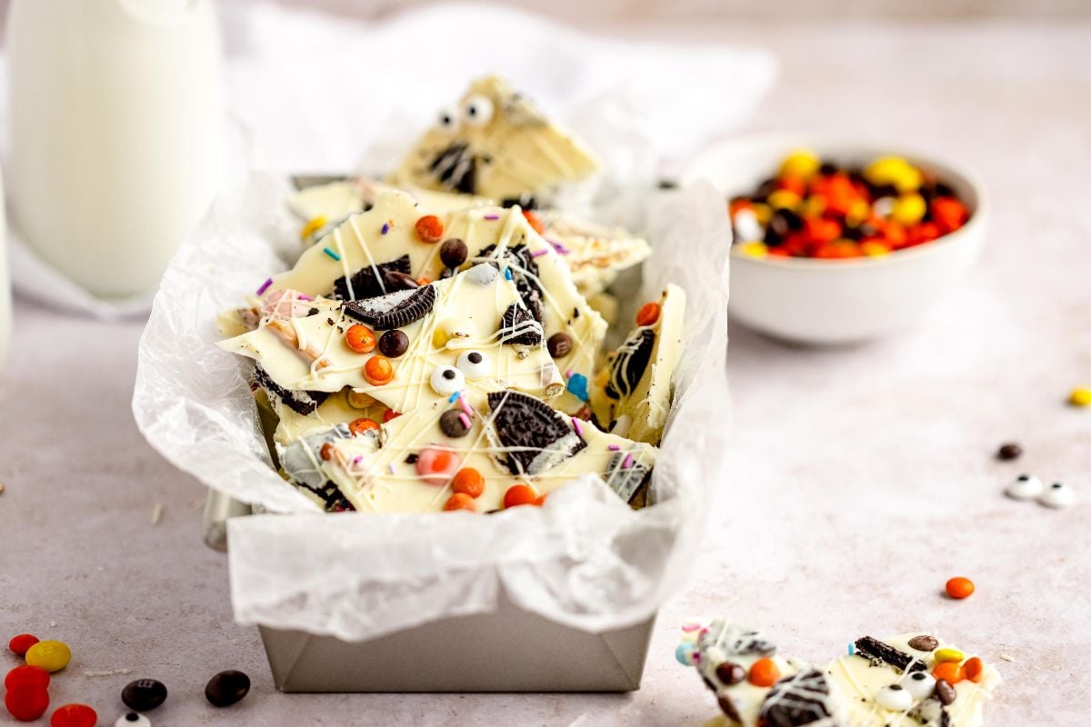 Halloween White Chocolate Candy Bark with googly monster eyes, sprinkles, and cookies sets in a parchment-lined pan; colorful candies scattered nearby. An easy candy bark recipe for a festive Halloween treat!.