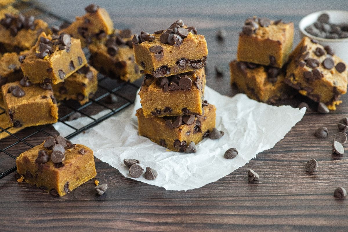 Squares of chocolate chip cookie dough fudge stacked on parchment paper and a cooling rack—proof that pumpkin and chocolate go together perfectly in every pumpkin squares with chocolate chips recipe.