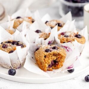 Blueberry muffins in white wrappers on a plate, paired with a cup of tea and scattered blueberries—this healthy blueberry muffin with carrots is a delightful twist inspired by classic carrot cake muffins.
