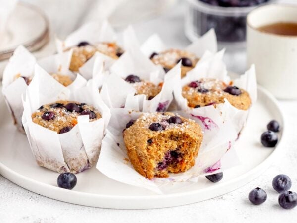 Blueberry muffins in white wrappers on a plate, paired with a cup of tea and scattered blueberries—this healthy blueberry muffin with carrots is a delightful twist inspired by classic carrot cake muffins.