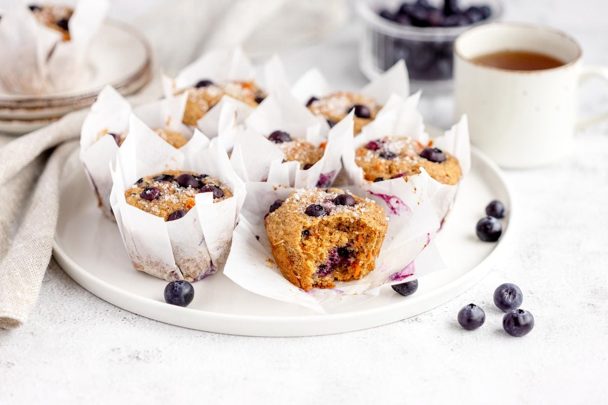 Blueberry muffins on a white plate, made using a carrot and blueberry recipe, served with a cup of tea and fresh blueberries scattered around.