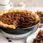 A pecan pie with a slice missing, surrounded by pecans, chocolate pieces, and coffee cups—enjoy a gooey pecan and chocolate dessert reminiscent of the best chocolate pecan pie.