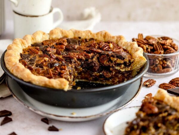 A pecan pie with a slice missing, surrounded by pecans, chocolate pieces, and coffee cups—enjoy a gooey pecan and chocolate dessert reminiscent of the best chocolate pecan pie.