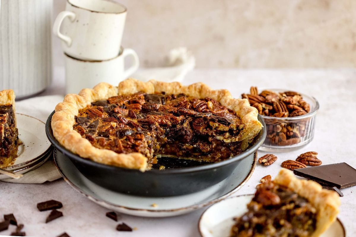 A gooey pecan and chocolate dessert with a slice missing, surrounded by pecans, chocolate pieces, and stacked teacups—just like Cracker Barrel pecan pie.