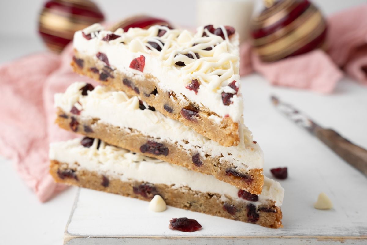Three stacked homemade bliss bars with white chocolate and cranberries, topped with icing, rest on a white board beside a pink cloth—an easy cranberry bar you’ll love to make at home.