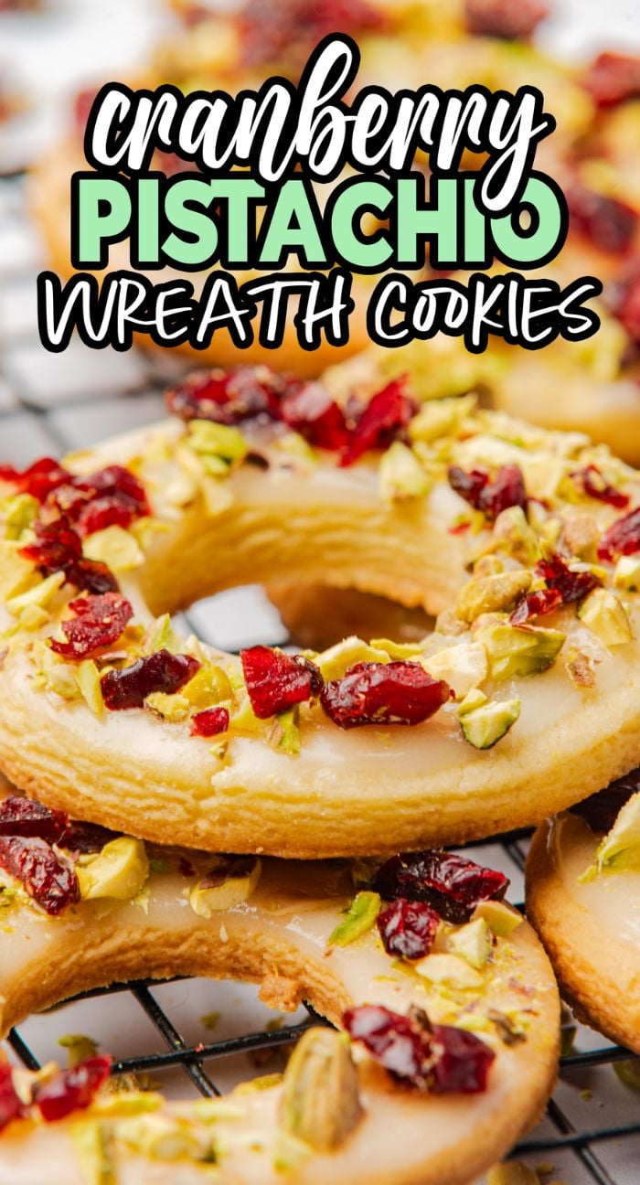 Cranberry pistachio wreath cookies are round treats topped with white icing, chopped pistachios, and dried cranberries on a cooling rack.