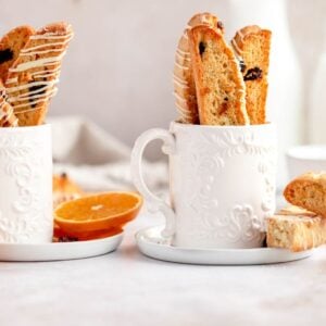 Two ornate white mugs filled with cranberry orange biscotti, surrounded by orange slices and a bottle of milk.