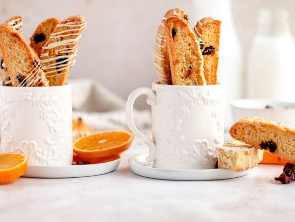 Two ornate white mugs filled with cranberry orange biscotti, surrounded by orange slices and a bottle of milk.