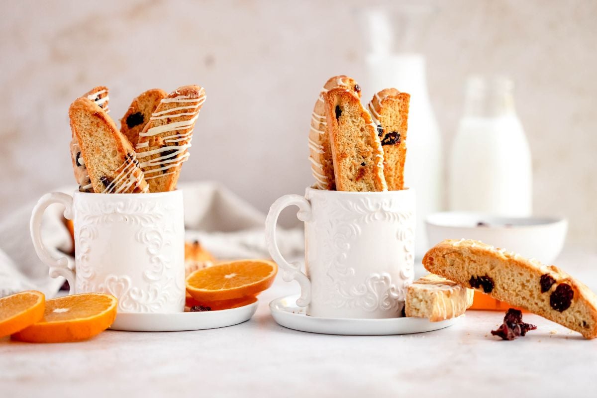 Two white mugs filled with cranberry orange biscotti, surrounded by orange slices and a bottle of milk in the background.