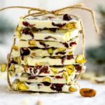 Stack of white chocolate bark, like almond bark, with pistachios and dried cranberries, tied with twine against a festive background—an easy holiday treat recipe perfect for gifting.