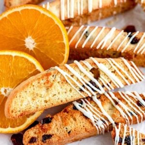 Cranberry orange biscotti with white drizzle and raisins are arranged next to fresh orange slices on a tray.