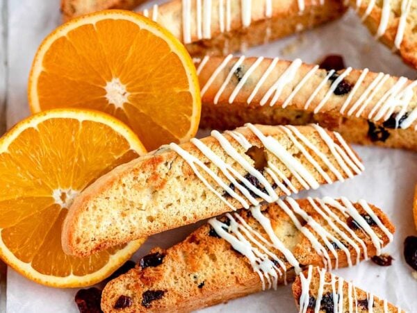 Cranberry orange biscotti with white drizzle and raisins are arranged next to fresh orange slices on a tray.