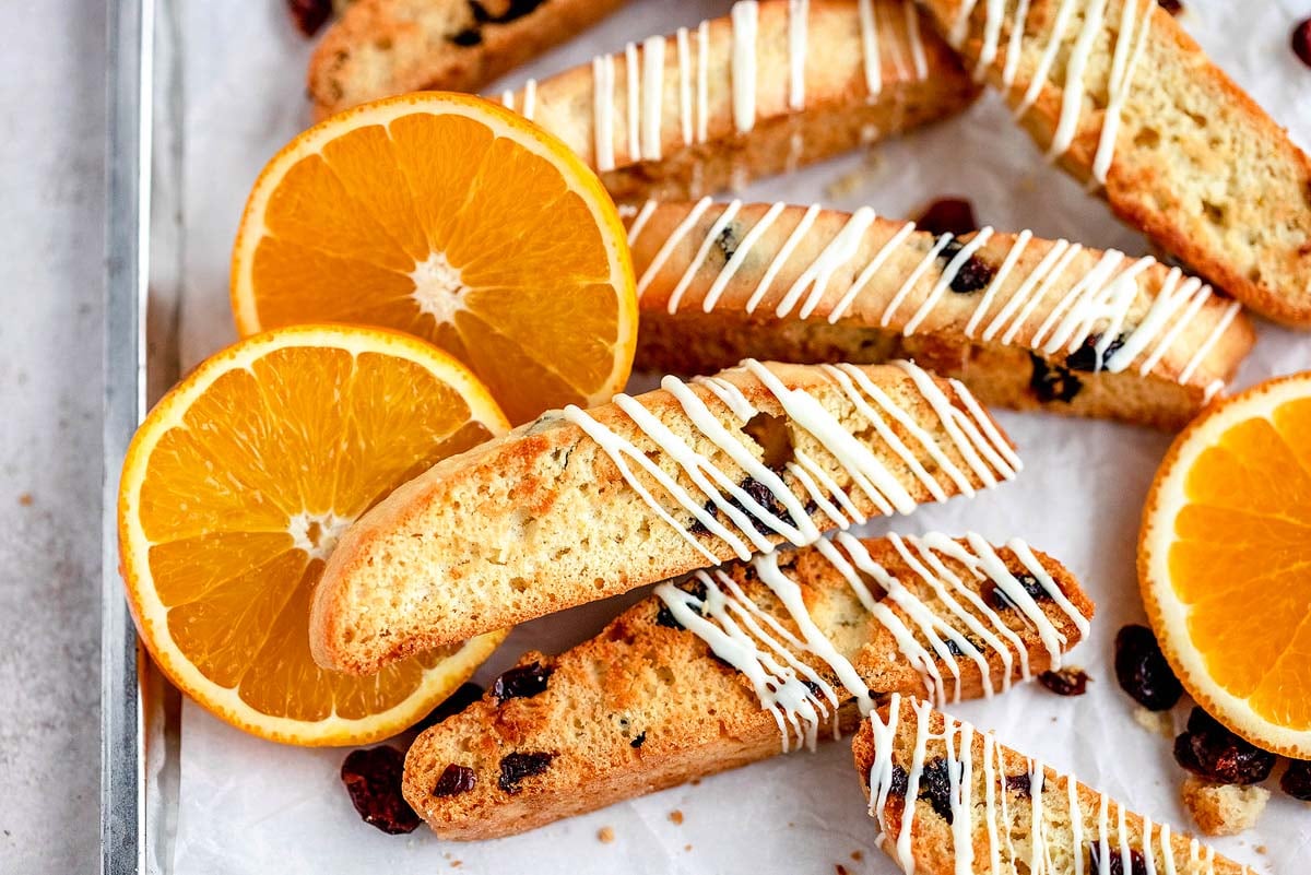 Cranberry orange biscotti with dried fruit and orange slices, drizzled with white icing, on a parchment-lined tray.