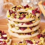 A stack of cranberry pistachio wreath cookies topped with white icing, garnished with pistachios and dried cranberries, elegantly arranged on a wooden board.