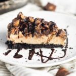 A slice of peanut butter cheesecake with chocolate drizzle and chopped candies on a chocolate graham crust, reminiscent of a decadent chocolate peanut butter pie.