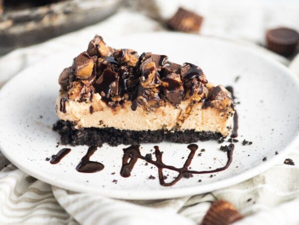 A slice of peanut butter cheesecake with chocolate drizzle and chopped candies on a chocolate graham crust, reminiscent of a decadent chocolate peanut butter pie.