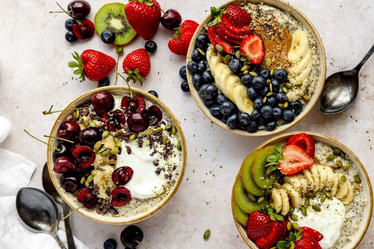 Three bowls of healthy oatmeal breakfast are topped with fresh fruit, seeds, and nuts, surrounded by spoons and assorted fruit—perfect for an oatmeal breakfast bowl to start your day.