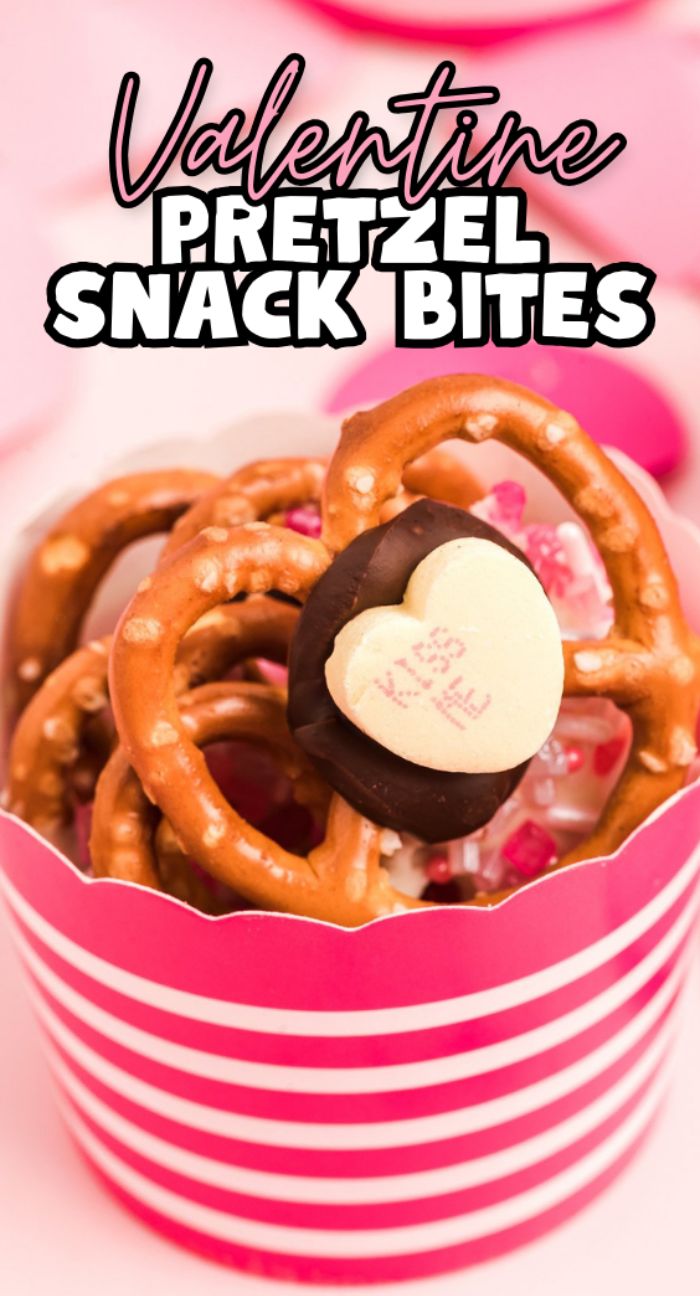 A striped pink cup holds Valentine Pretzel Snack Bites—pretzels topped with chocolate and a heart-shaped candy—for a festive Valentine’s Day treat.