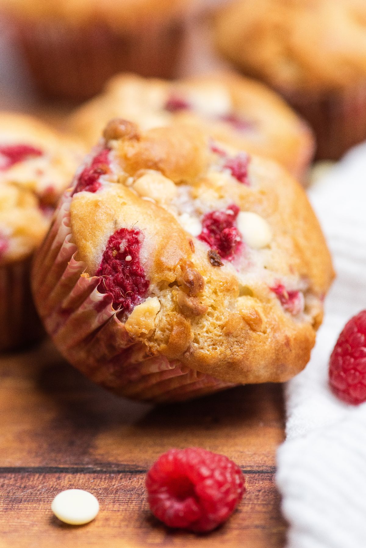A raspberry white chocolate muffin sits on a wooden surface, surrounded by fresh raspberries and a white cloth—an inviting scene for lovers of raspberry muffins.