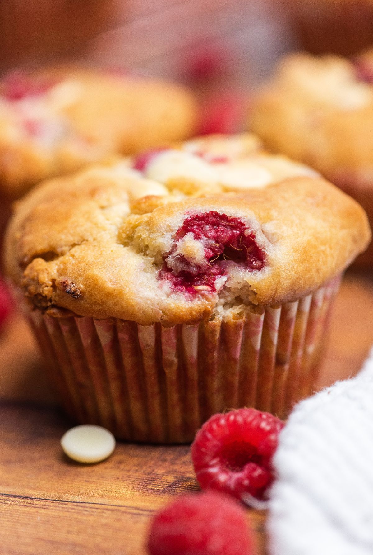 A raspberry white chocolate muffin with a bite taken out, topped with white chocolate chips, sits on a wooden surface.