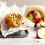 A muffin with a bite taken out sits in a muffin tin, with apples and slices in the background—perfect for showcasing your favorite apple crumble muffins recipe.