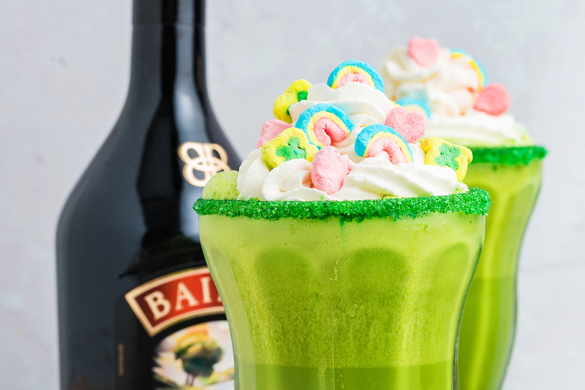 A green Irish cream milkshake topped with whipped cream and colorful cereal sits beside a bottle of Baileys Irish Cream.