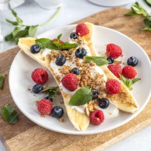 Breakfast Banana Split with yogurt, granola, banana and fresh berries—raspberries, blueberries, and mint—on a white plate. A fun alternative to a smoothie bowl for a refreshing start to your day.