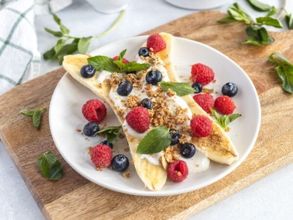 Breakfast Banana Split with yogurt, granola, banana and fresh berries—raspberries, blueberries, and mint—on a white plate. A fun alternative to a smoothie bowl for a refreshing start to your day.