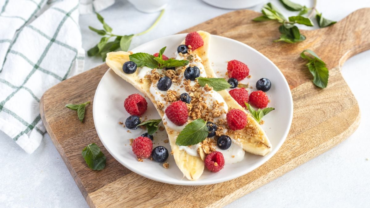 Breakfast Banana Split with yogurt, granola, banana and fresh berries—raspberries, blueberries, and mint—on a white plate. A fun alternative to a smoothie bowl for a refreshing start to your day.